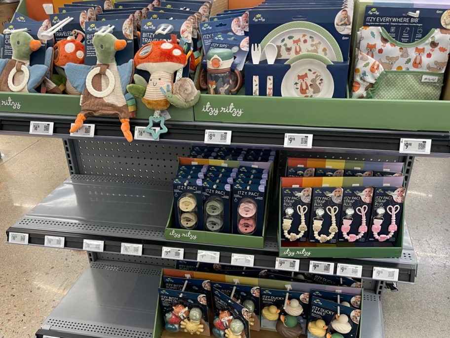 A retail store display shelf stocked with Itzy Ritzy baby products, including plush toys, dish sets, pacifiers, pacifier clips, and bath toys.