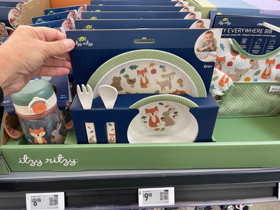 A hand holding a packaged Itzy Ritzy children's dishware set featuring plates and utensils with woodland animal illustrations, displayed on a store shelf alongside a sippy cup and bib.