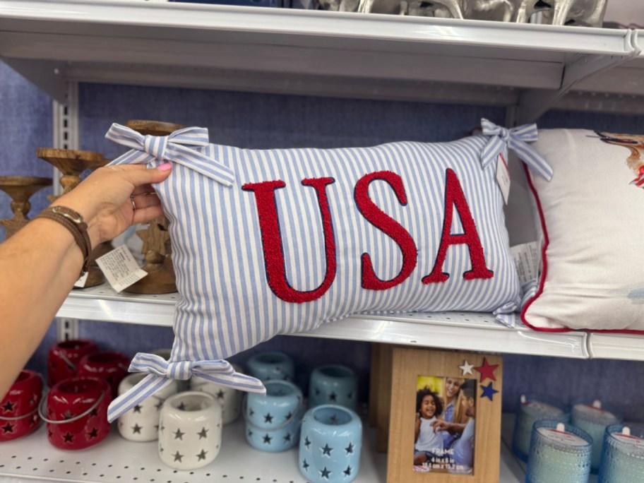 A hand is holding a rectangular pillow with "USA" in bold red letters on a blue and white striped background. The pillow is adorned with four corner bows.