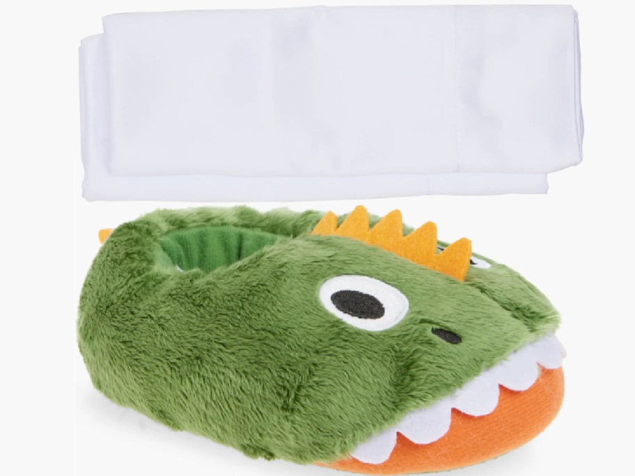 Green dinosaur-themed slipper with fluffy texture, featuring white teeth, and orange details. White sham set in the background.