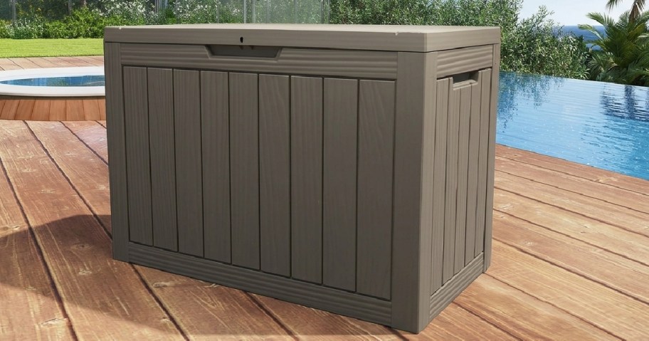 A large, brown outdoor storage box on a wooden deck beside a pool, surrounded by greenery.