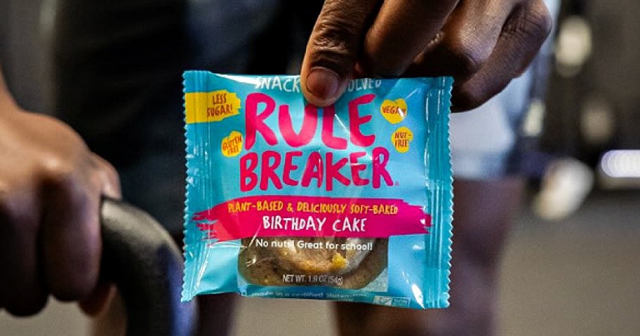Person holding a brightly colored "Rule Breaker" snack package with birthday cake flavor. The packaging highlights it's plant-based and nut-free.