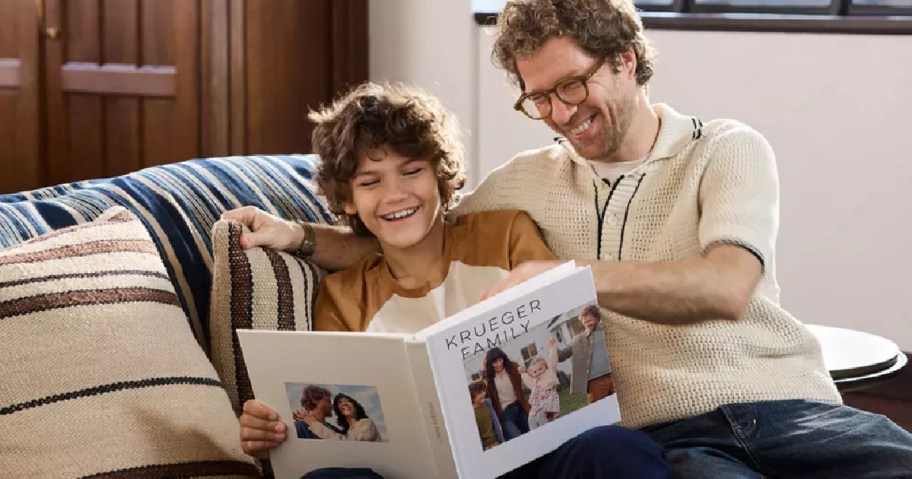 A man and a boy are sitting on a couch, looking at a photo book titled "Krueger Family." Both are smiling and enjoying the moment together.