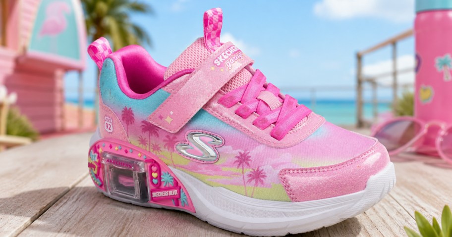 Pink and blue ombre Skechers children's sneakers with palm tree graphics on a wooden surface with a beach background.