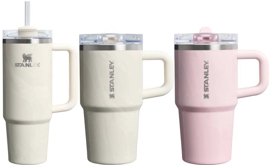 Three Stanley travel mugs in different colors: a white mug with a straw, a beige mug, and a pink mug.