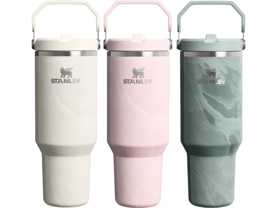 Three Stanley insulated tumblers in various colors: a white one, a pink one, and a gray marbled one, all with handles.