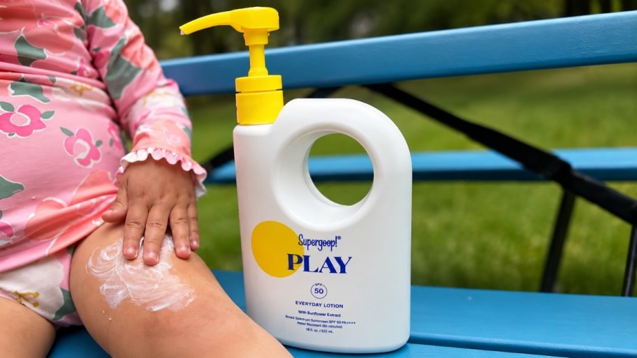 a baby applying sunscreen sitting next to a jug of sunscreen on a bench