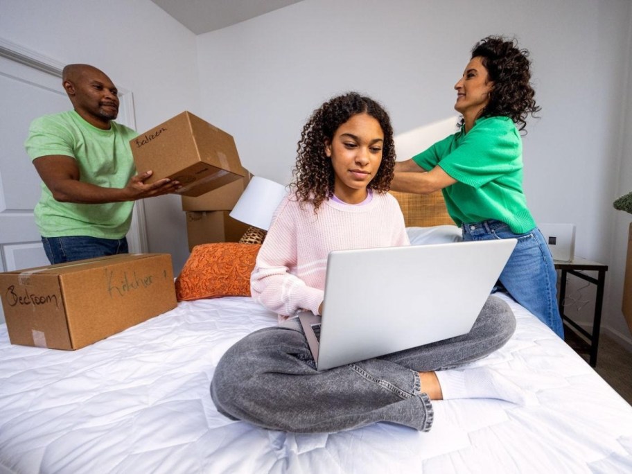 a woman sitting on a bed using a laptop with people unpacking boxes behind her