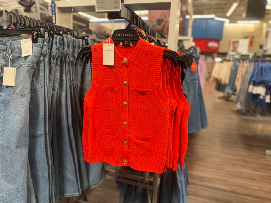 A vibrant orange knit vest with gold buttons hangs on a rack beside rows of denim jeans in a clothing store.