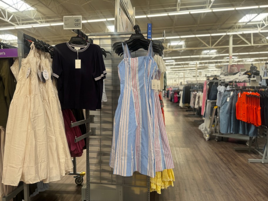 A clothing rack displays a black shirt and a pastel striped dress in a well-lit store aisle, with more clothes visible in the background.