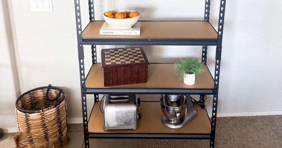 A metal shelving unit with three shelves. The top shelf holds a bowl of oranges and a book. The middle shelf has a wooden chess set and a small potted plant. The bottom shelf contains a toaster and a mixing machine. A woven basket is positioned on the left side of the unit.