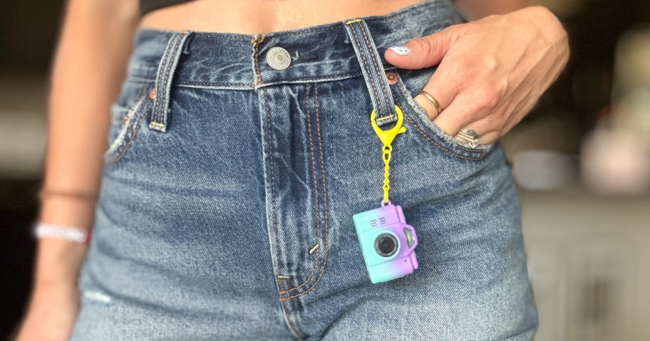 A close-up of a person's midsection wearing high-waisted denim shorts, with a mini camera keychain attached to the pocket.
