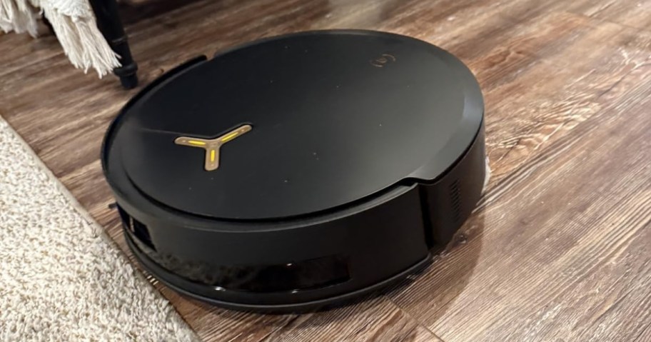 Black robot vacuum cleaner on a wood floor next to a rug. 