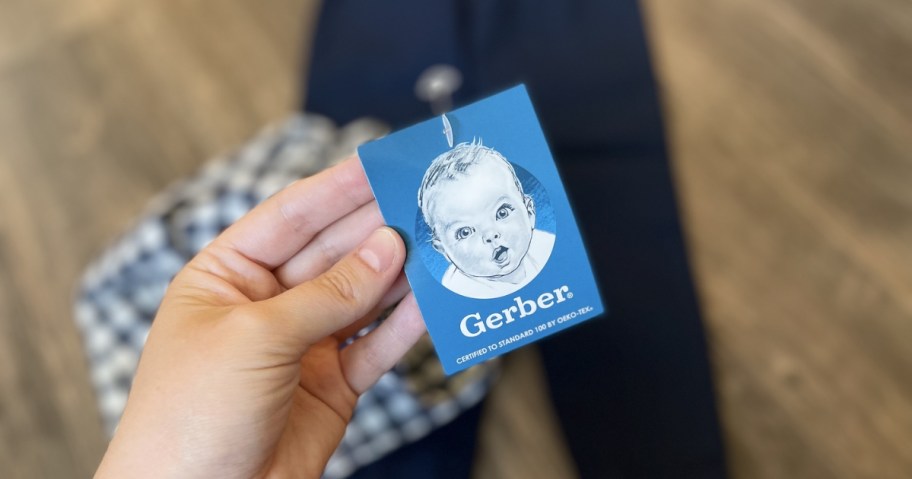 person holding gerber baby tag with logo and baby clothes on floor in background