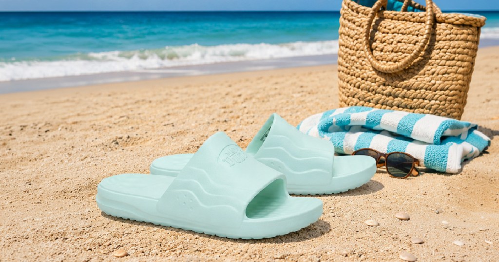 Light green hey dudes slides on sand next to beach bag and towel