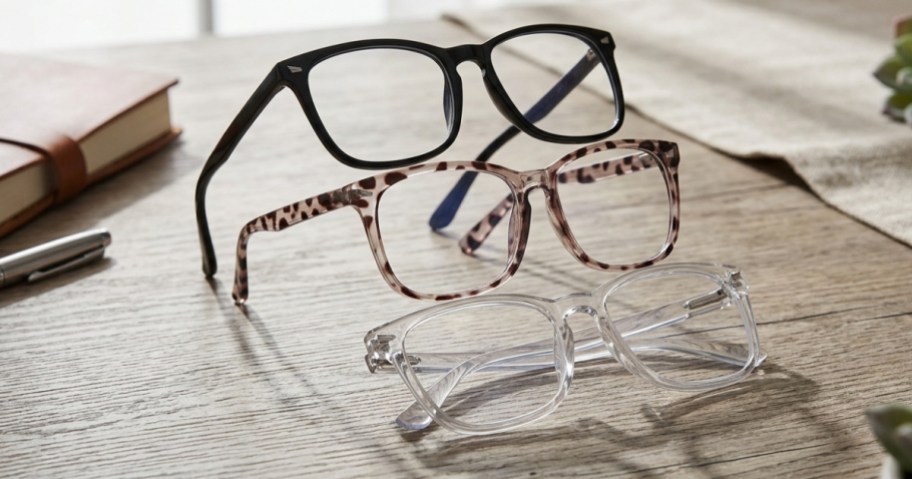 A stack of three pairs of glasses on a wooden surface, with a notebook and pen in the background.