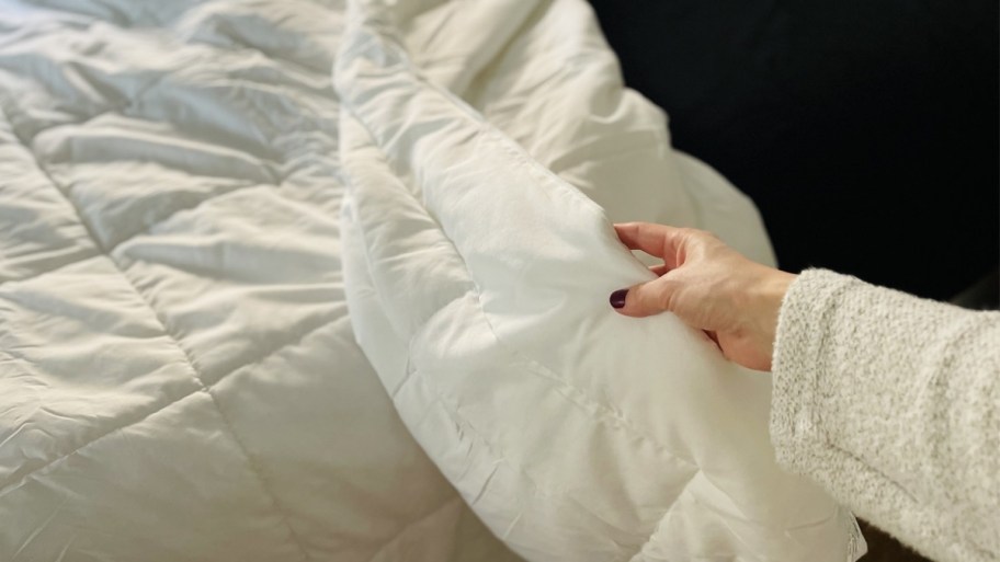 a womans hand turning back the edge of a white comforter on a bed 