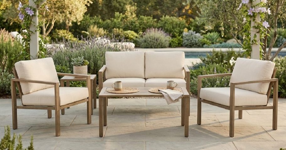 A cozy outdoor seating area with two armchairs and a loveseat, all featuring beige cushions. A wooden table sits between the seats, complemented by a small potted plant and some decorative items. The setting is surrounded by lush greenery and flowers under a pergola.