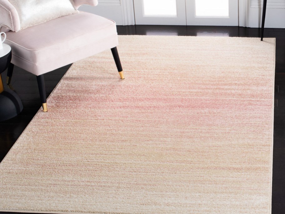 A rectangular area rug with a subtle cream to soft pink ombre pattern, placed on a dark wood floor partially underneath a pale pink chair.