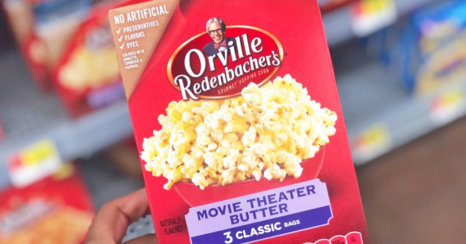 hand holding a red box of Orville Redenbacher’s Movie Theater Butter Popcorn in store aisle