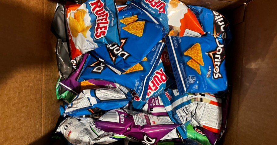 An open cardboard box filled with an assortment of single-serve Ruffles and Doritos chip bags.