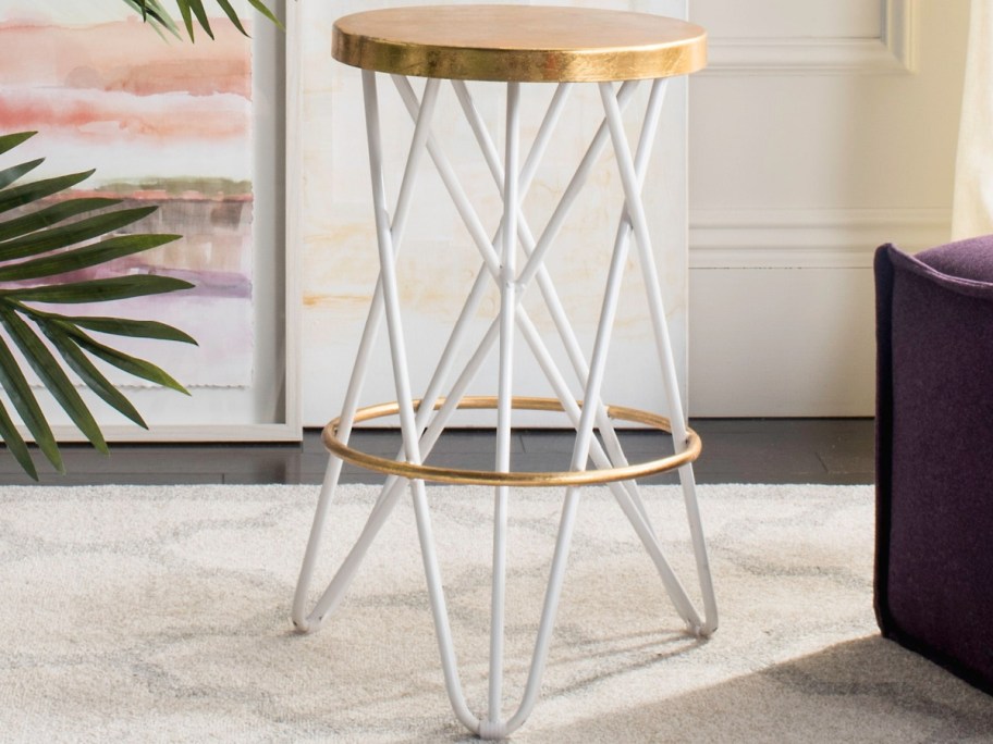 A counter-height metal bar stool with a round gold top, white crisscrossing geometric legs, and a gold footrest ring on a rug.