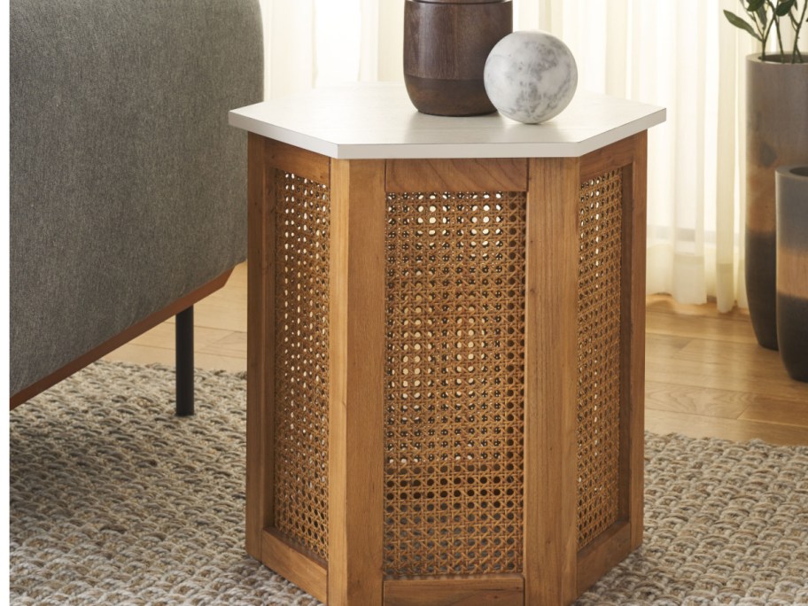 A hexagonal wooden accent table with a white top and woven cane panel sides next to a grey sofa.