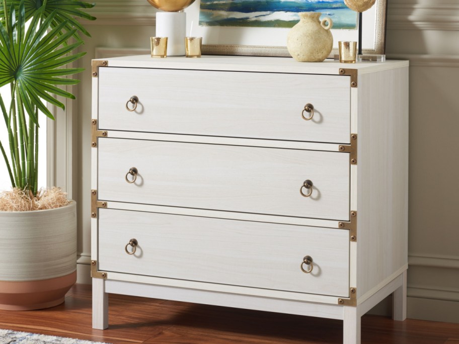 A bleached white three-drawer chest featuring brass corner hardware and ring pulls in a decorated room.