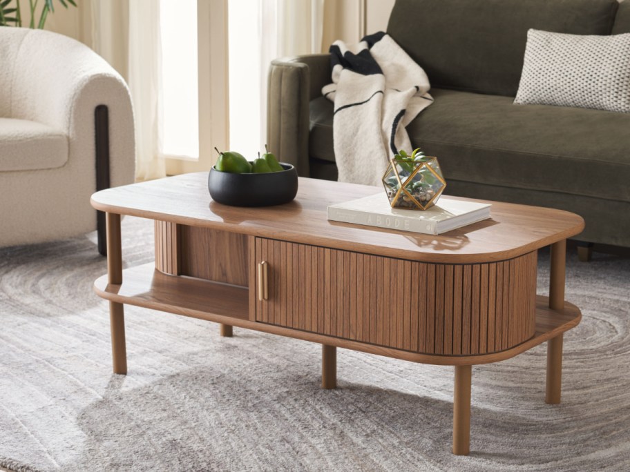 A modern wooden coffee table with a fluted tambour door cabinet, lower shelf, and tapered legs in a styled living room.