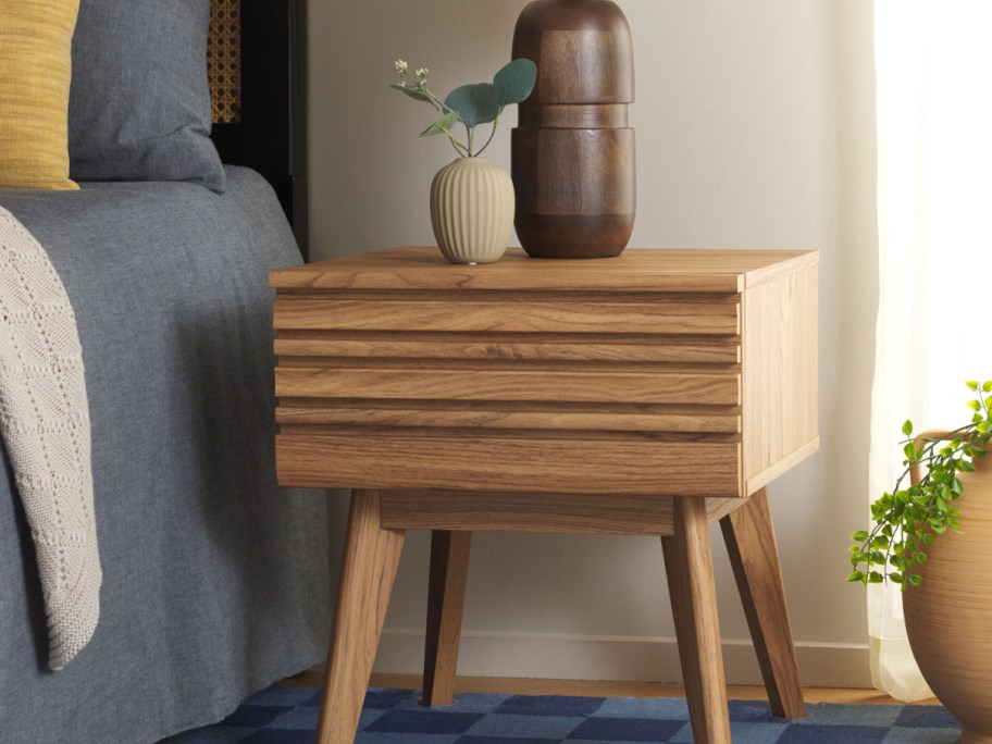 A wooden nightstand with horizontal slat drawer fronts and angled legs positioned next to a made bed.