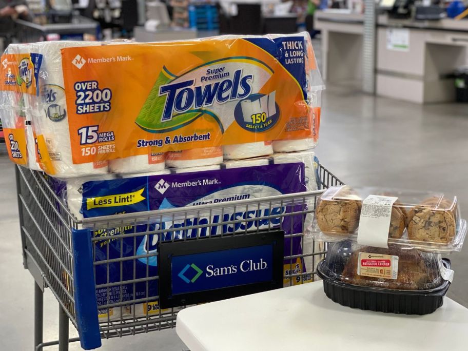 A Sam's Club shopping cart with Member's Mark toilet paper and paper towels in it next to a table with a pack of cookies and a rotisserie chicken on it