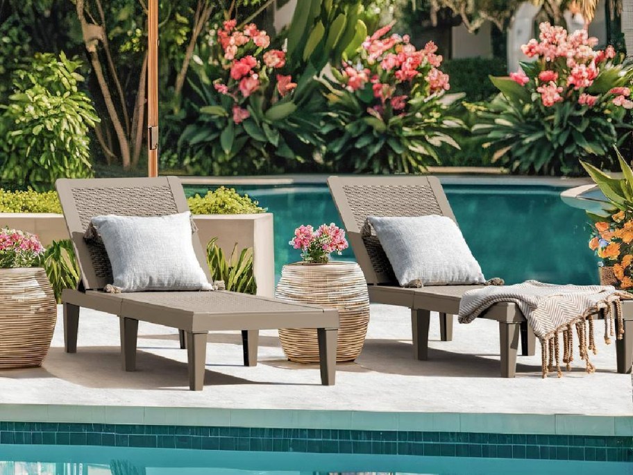 Two lounge chairs with cushions beside a swimming pool, surrounded by lush greenery and colorful flowers.
