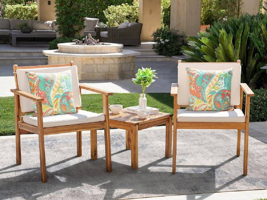 A patio setting with two wooden chairs, each with colorful patterned cushions, and a small table between them, surrounded by greenery and a water feature in the background.
