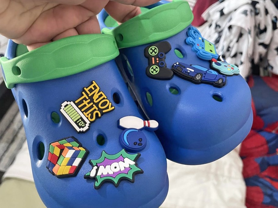 hand holding up a pair of blue and green clogs with charms all over them