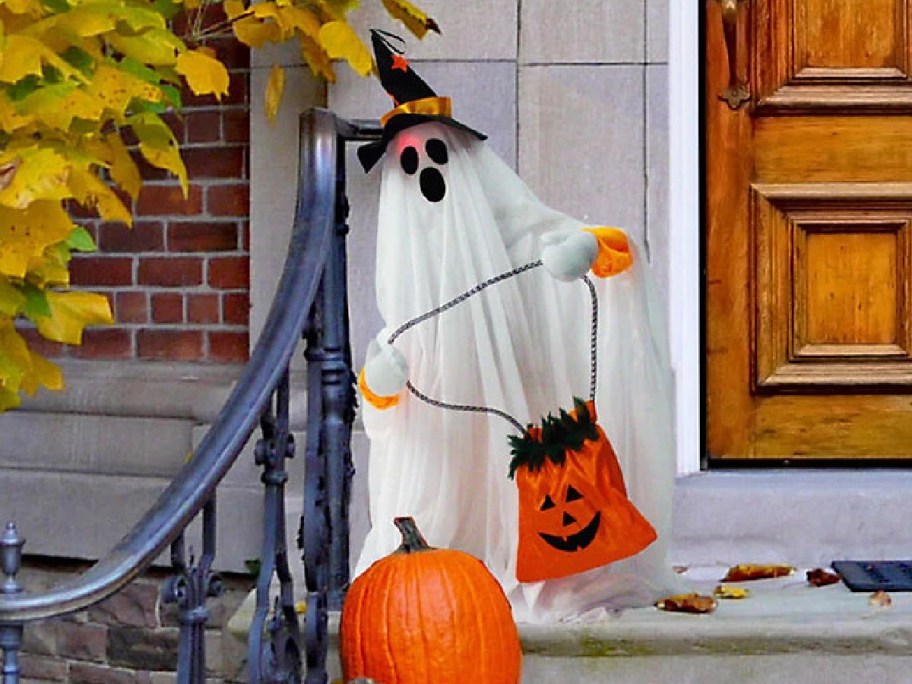 A white ghost decoration wearing a black witch hat and holding an orange jack-o'-lantern trick-or-treat bag, standing on porch steps next to a real pumpkin.