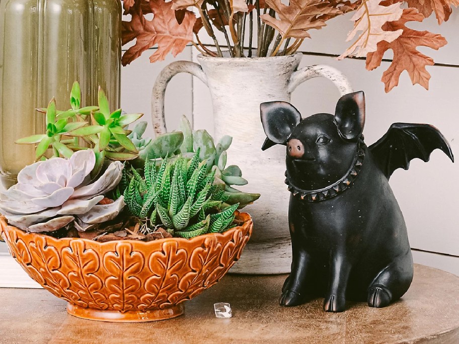 A black statue of a pig with bat wings wearing a studded collar sitting next to an orange, leaf-patterned bowl filled with succulents.