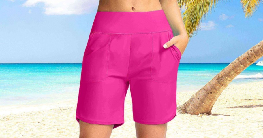 A person wearing bright pink high-waisted shorts with front pockets, standing on a sandy beach with the ocean and a palm tree trunk in the background.