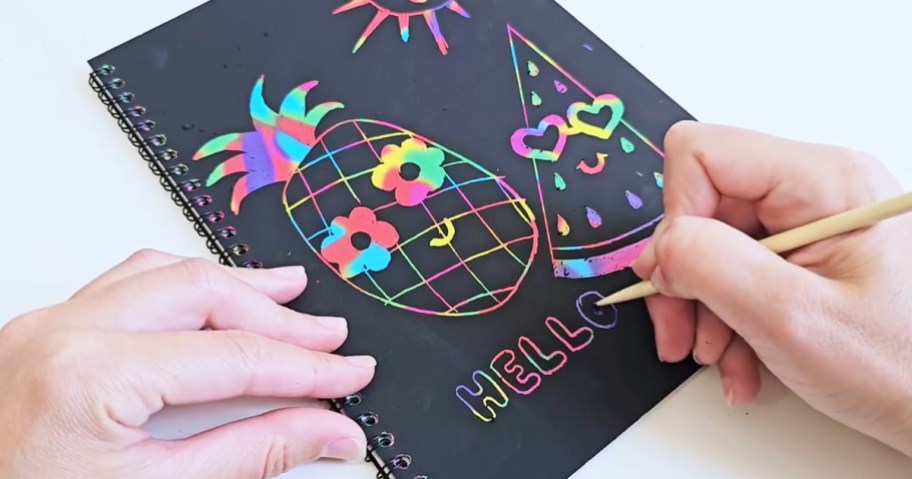 person drawing pineapple and watermelon doodles on rainbow scratch art paper