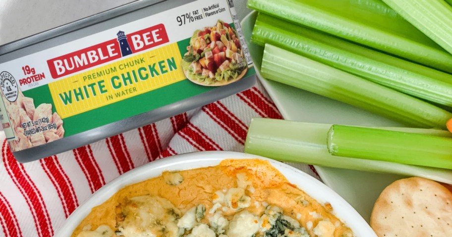 A can of Bumble Bee Premium Chunk White Chicken next to a bowl of baked chicken dip topped with blue cheese, served with celery sticks and crackers on a red and white striped towel.
