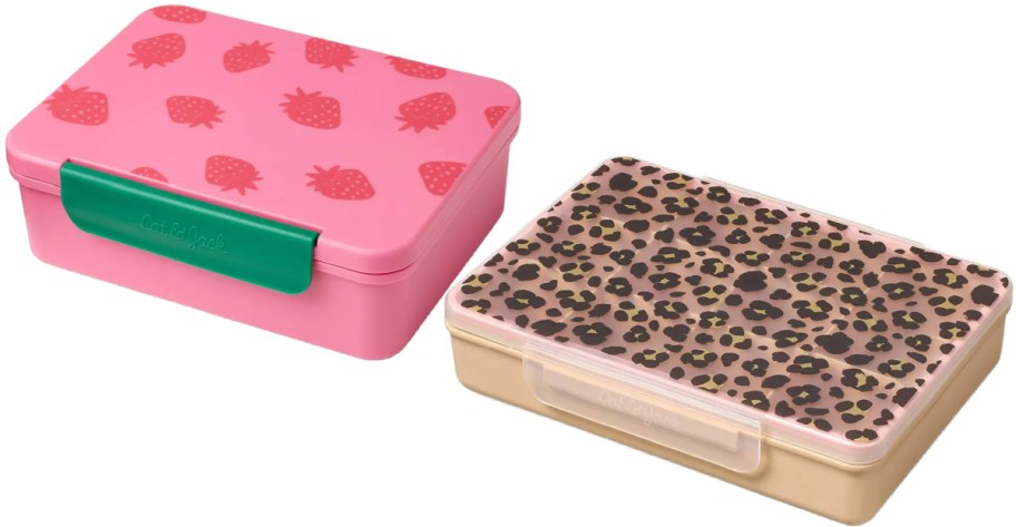 Two bento lunch boxes side-by-side: a pink one with a strawberry pattern and a green latch, and a tan one with a leopard print lid and a clear latch.