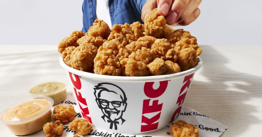 A large KFC bucket filled with popcorn chicken sitting on a light wooden table next to two small containers of dipping sauce, with a person's hand reaching in to grab a piece.