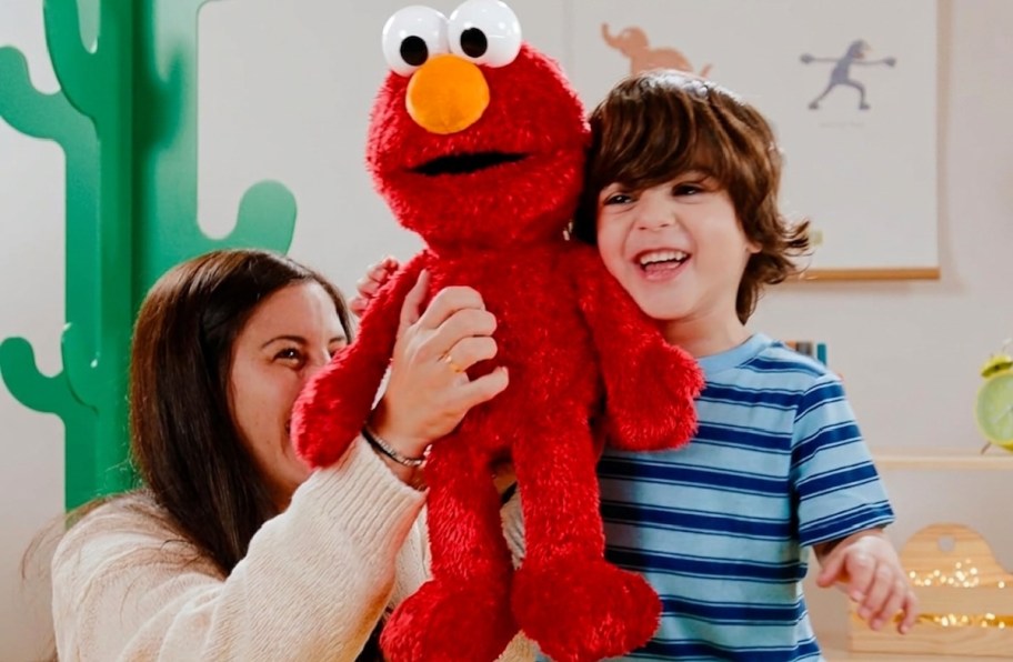 A child holds a large red Tickle Me Elmo doll, smiling while a woman beside him supports the doll.
