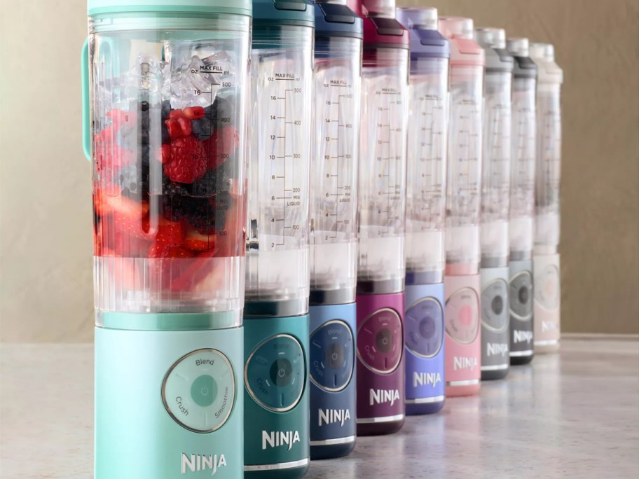 There's a row of colorful Ninja blenders and clear containers filled with fruit and ice.