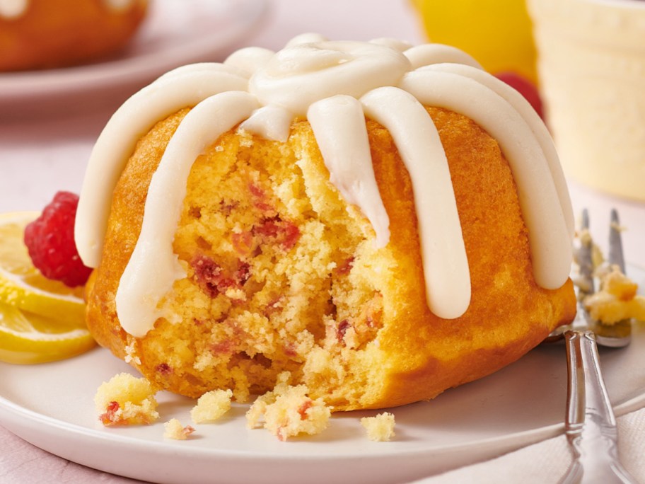 A small yellow bundt cake speckled with raspberry pieces and topped with thick white frosting, with a piece taken out to reveal the crumb inside, sitting on a plate garnished with lemon slices and a fresh raspberry.