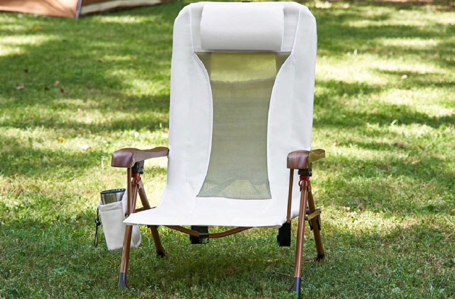 A white folding outdoor chair with a mesh back panel, wooden armrests, and an attached side cup holder sitting on green grass.