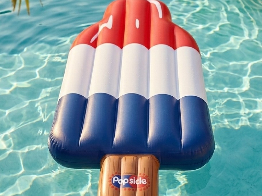 A large inflatable popsicle-shaped float in a swimming pool. The float is designed with red, white, and blue colors.