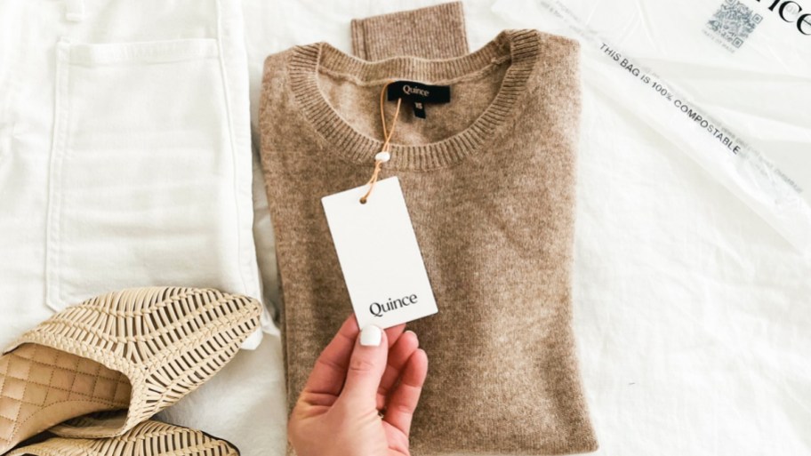 a womans hand holding a cardboard quince tag attached to a sweater