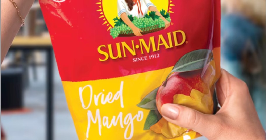 A person holding a bag of Sun-Maid Dried Mango, featuring the brand logo and an image of mangoes on the packaging.