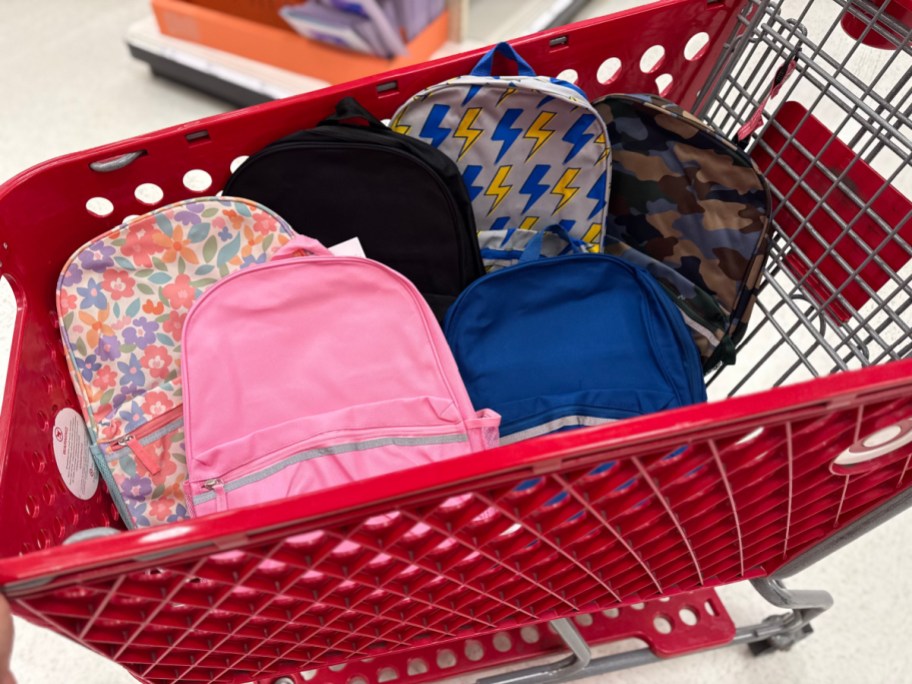 A red Target shopping cart filled with a variety of backpacks in solid pink, blue, and black along with floral, lightning bolt, and camouflage patterns.