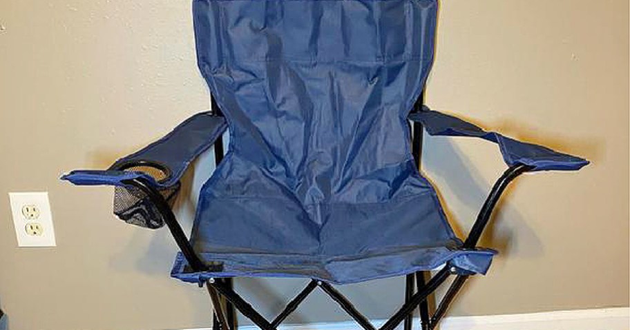 A dark blue folding camping chair with a black frame and a mesh cup holder in the armrest, positioned indoors against a beige wall.